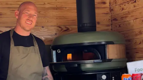 Dan Puddle Dan Puddle in his mobile pizza van with a small pizza-oven in the background. He wears an apron and is smiling.