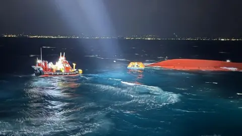 South Korean Coastguard The orange upturned hull of a boat is visible in the water. It's nighttime and the waves are illuminated by lights. People are seen in a small lifeboat, plus another boat nearby.