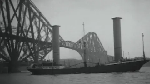 Pathe News Black and white image of the Buckau, with a black hull and two large towers sticking up from the deck. It has just passed under the iconic Forth Bridge
