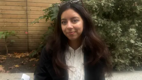 Martina is smiling. She has long, dark brown hair, and is wearing a white knitted zipped hoodie under a black jacket.