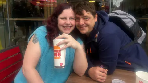 Joanne, left, smiling, wearing a sleeveless blue top with shoulder length wavy hair, holding a take-out beverage cup. Chris, right, smiles, and is wearing a dark blue top and a rucksack. He is leaned over with his face next to Joanne's.