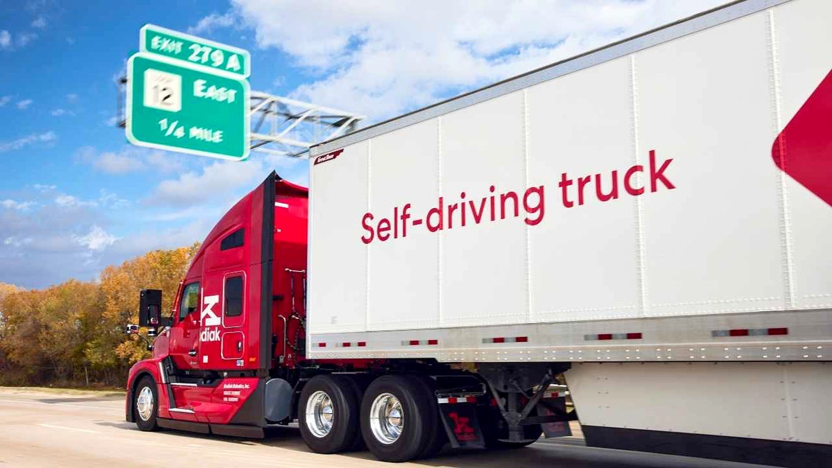 A red Kodiak self-driving semi-truck drives on a highway under a blue sky near an exit sign.